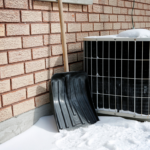 Snow-covered air conditioner unit outside a brick home with a snow shovel leaning nearby, illustrating winter HVAC maintenance considerations