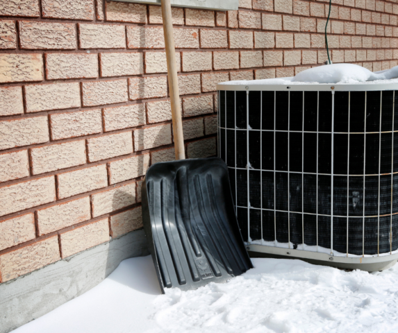 Snow-covered air conditioner unit outside a brick home with a snow shovel leaning nearby, illustrating winter HVAC maintenance considerations