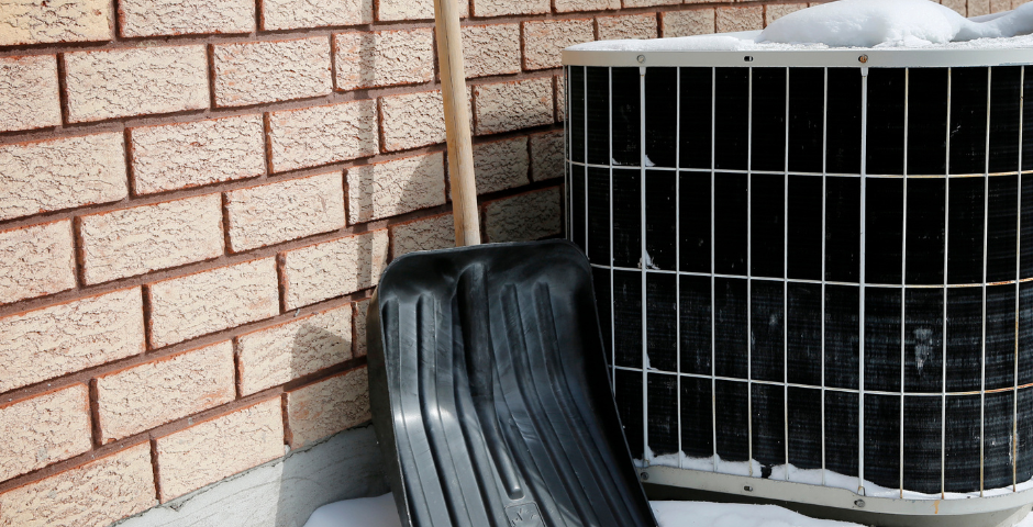 Snow-covered air conditioner unit outside a brick home with a snow shovel leaning nearby, illustrating winter HVAC maintenance considerations